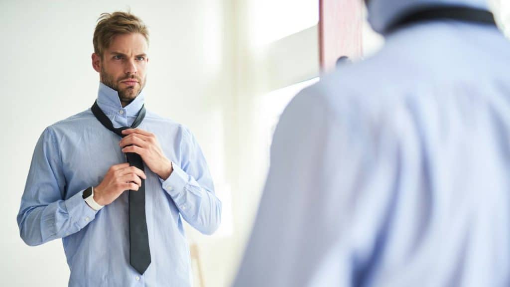 A serious man in a light blue shirt adjusts his black tie while looking at his reflection.
