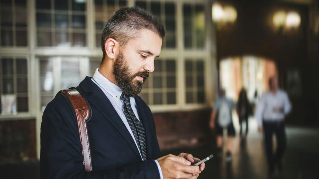 A focused businessman with a beard and shoulder bag looks down at his phone indoors.