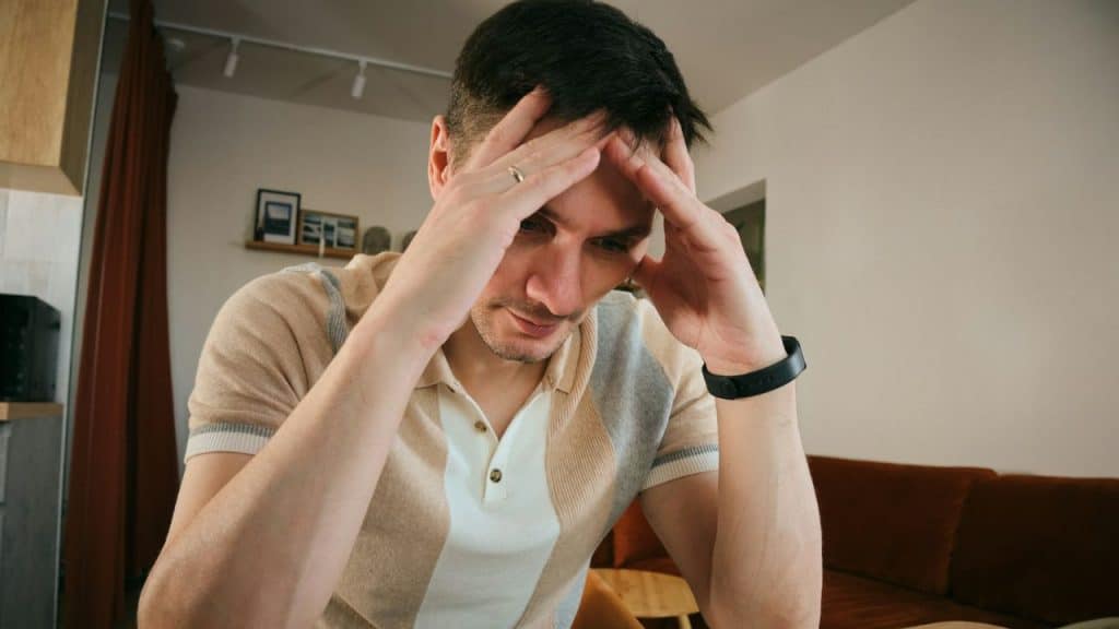 A worried man in a striped polo shirt sits with both hands pressed against his temples.