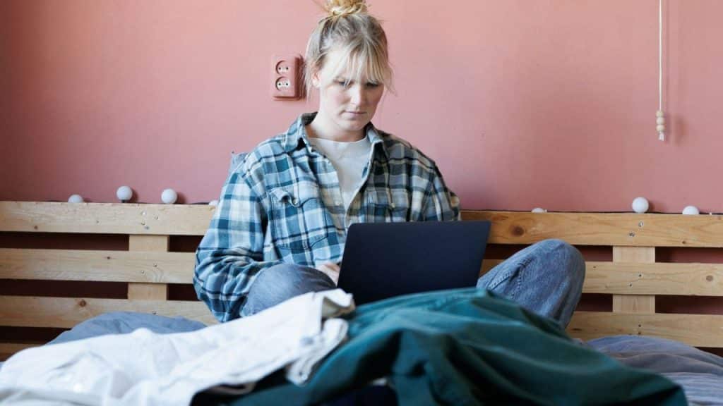 A serious blonde woman sits cross-legged on a bed, working on a laptop.
