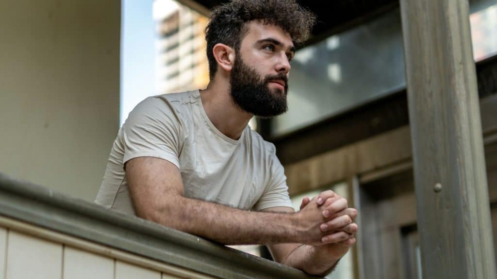 A serious, bearded man with curly hair leans on a railing, looking into the distance.