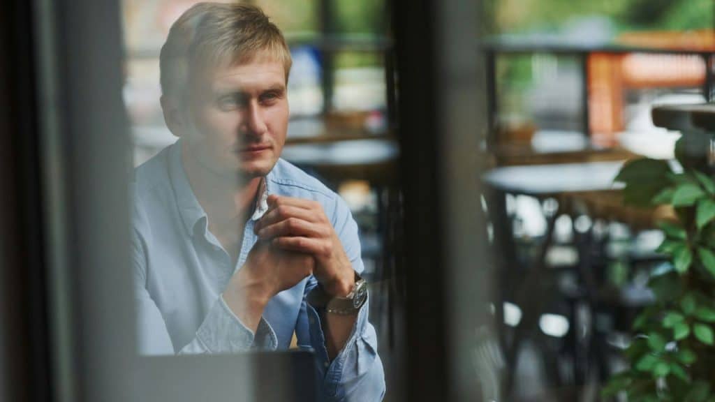 A man sits alone at an outdoor cafe table, resting his clasped hands.
