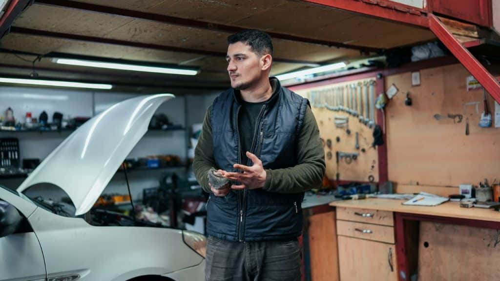 A male mechanic stands thoughtfully in a garage with a car's hood up.
