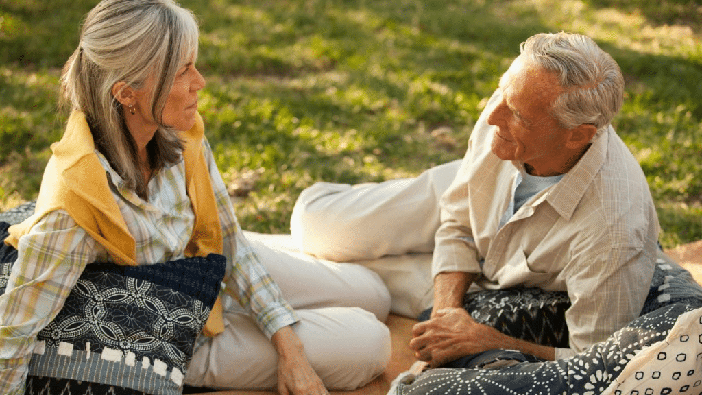 An elderly couple is relaxed, sitting on a blanket on the grass and having a conversation.