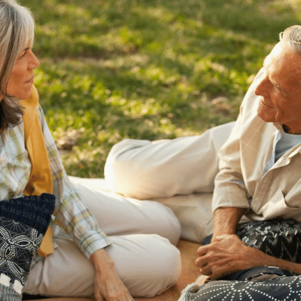 An elderly couple is relaxed, sitting on a blanket on the grass and having a conversation.