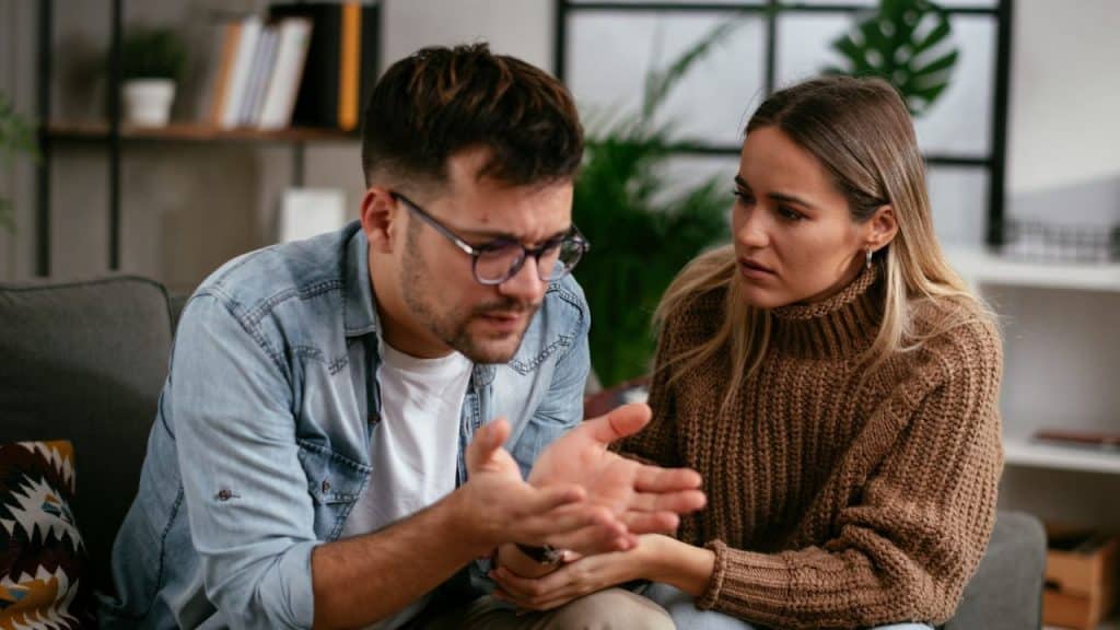 A serious couple sits on a couch discussing something with expressive hand gestures.