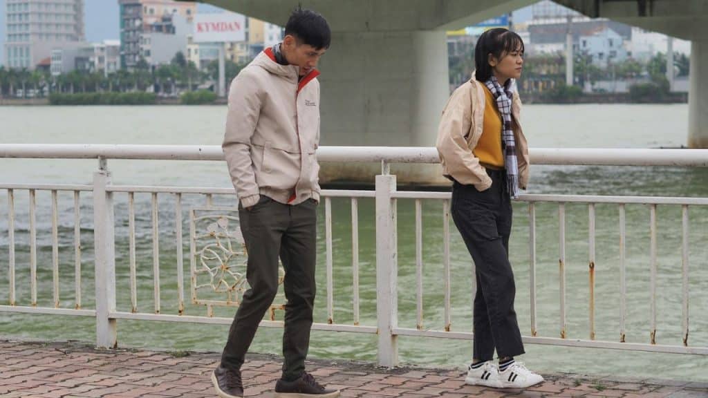 A young couple walking apart on a riverside promenade near a bridge.