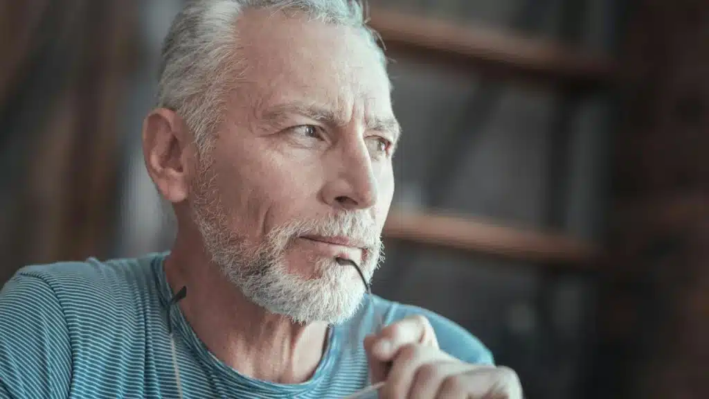A thoughtful senior man with a salt-and-pepper beard holds glasses near his mouth.