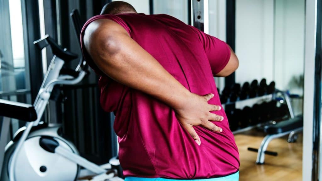 A man in a burgundy shirt is standing in a gym, clutching his lower back in pain.