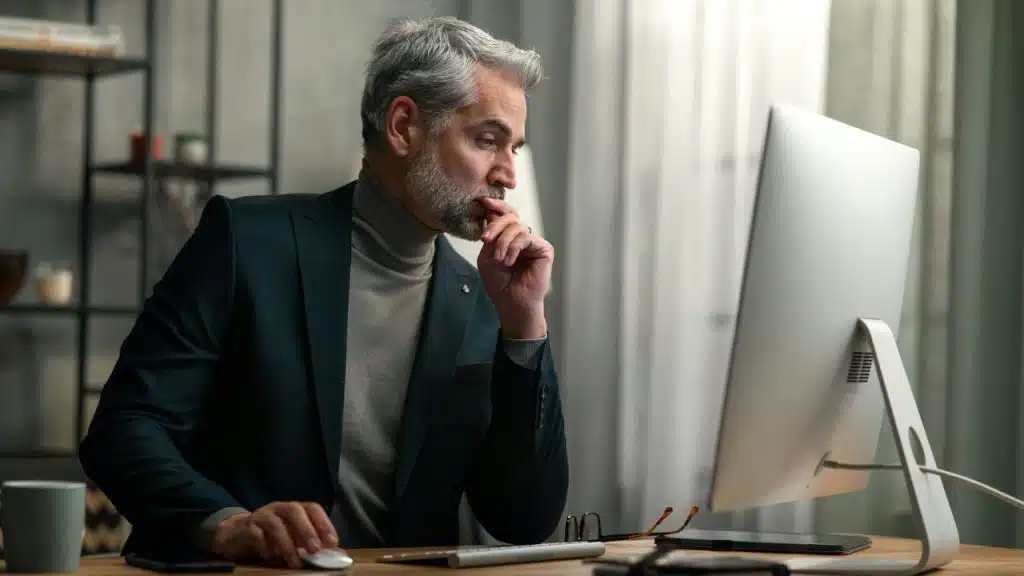 A gray-haired, bearded man in a suit jacket and turtleneck stares intently at a computer monitor.