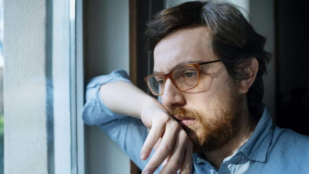 A worried man with a beard and glasses rests his head on his arm, looking out a window.