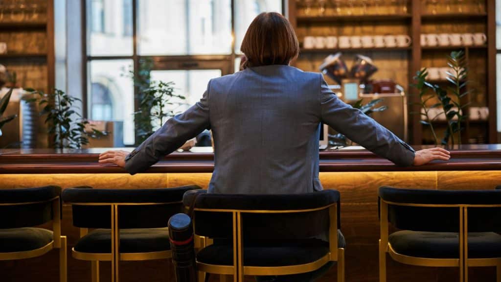 A person in a gray suit sits alone at a dark bar with their back to the camera.