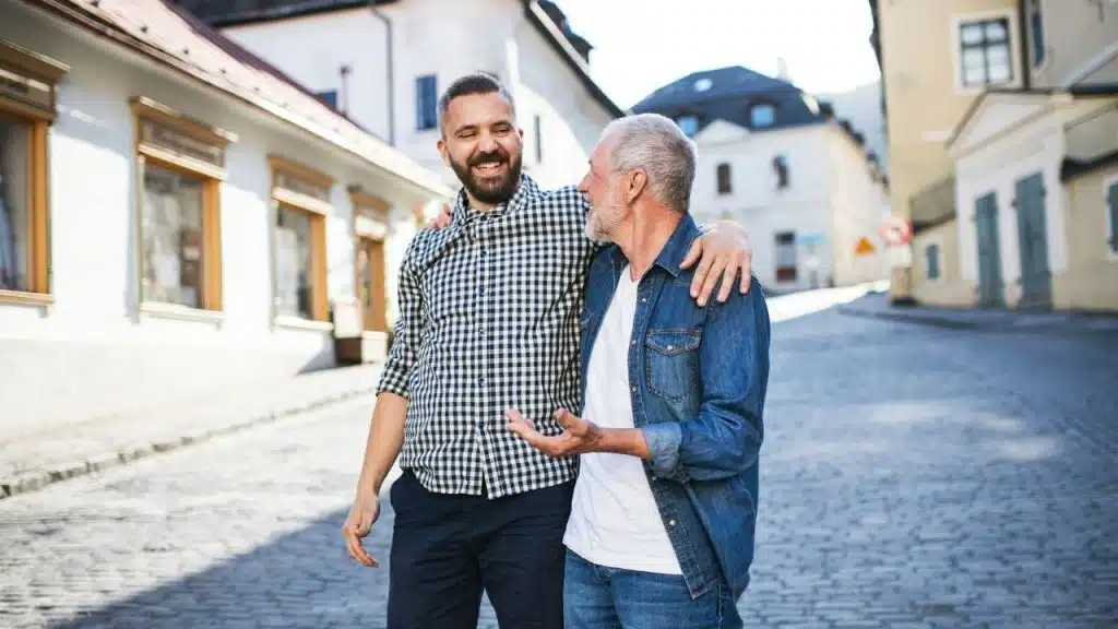 A happy middle-aged son with a beard walks arm-in-arm with his senior father on a cobblestone street.