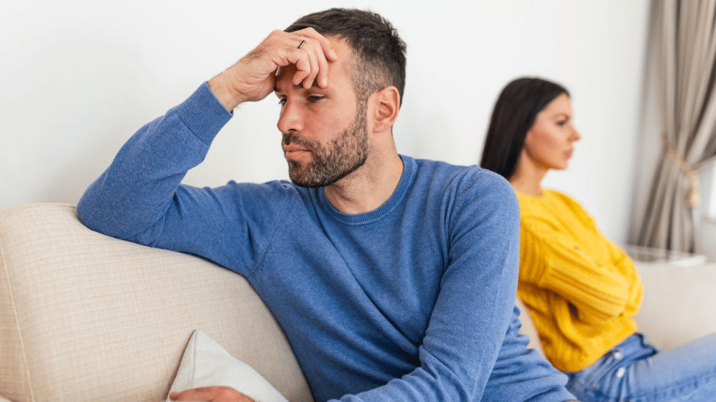 A stressed man with his hand on his forehead sits on a couch while a woman faces away.