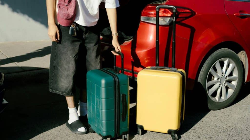 A person loads a green and a yellow rolling suitcase near the back of a red car.