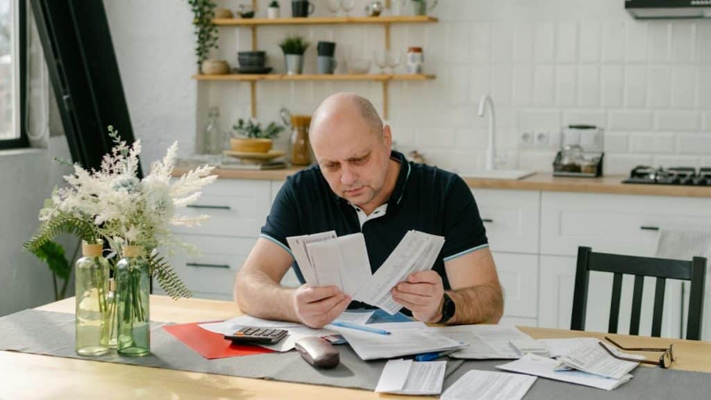 A serious bald man sits at a kitchen table, intently reviewing multiple bills.