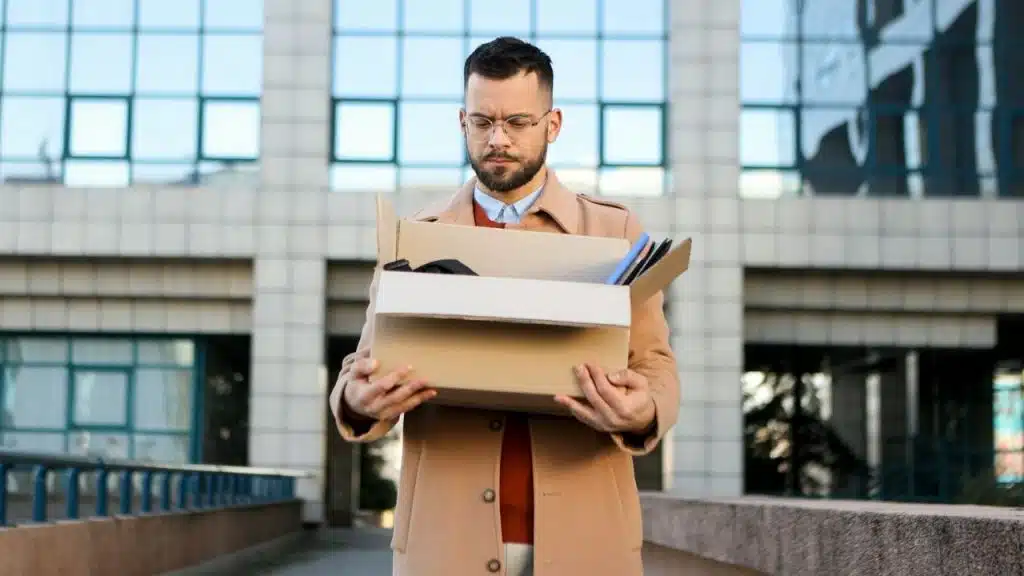 A distraught, bearded man in a trench coat holds a box of his belongings outside an office building.