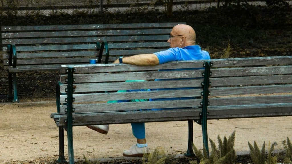 A solitary, bald senior man sits on a wooden park bench, looking away.