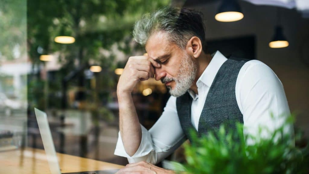 A middle-aged businessman with a beard sits at a laptop, resting his head in his hand in frustration.
