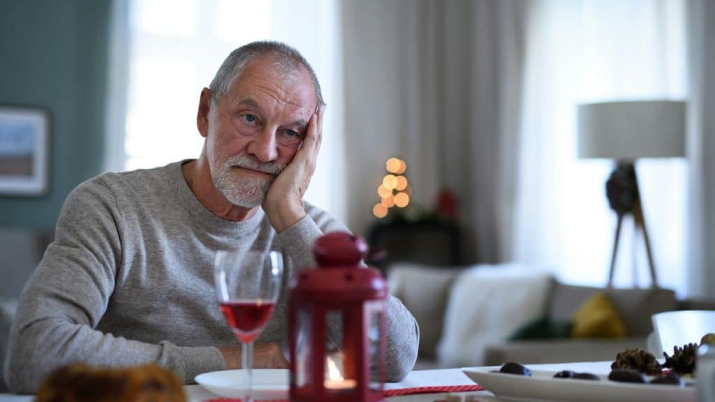 A glum, older man sits alone at a dinner table, resting his head on his hand.