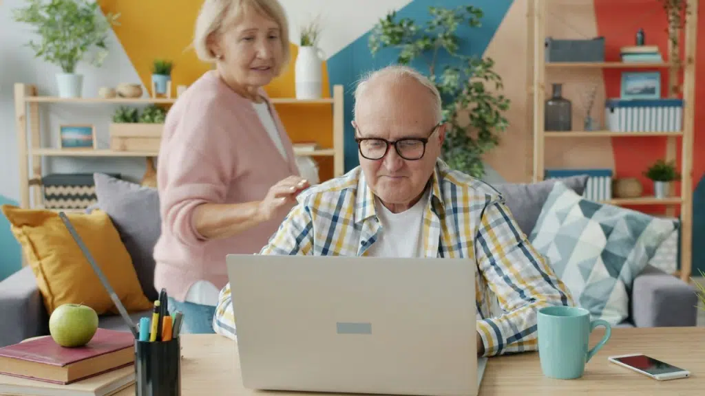 An older man works intently on a laptop while an older woman stands behind him.