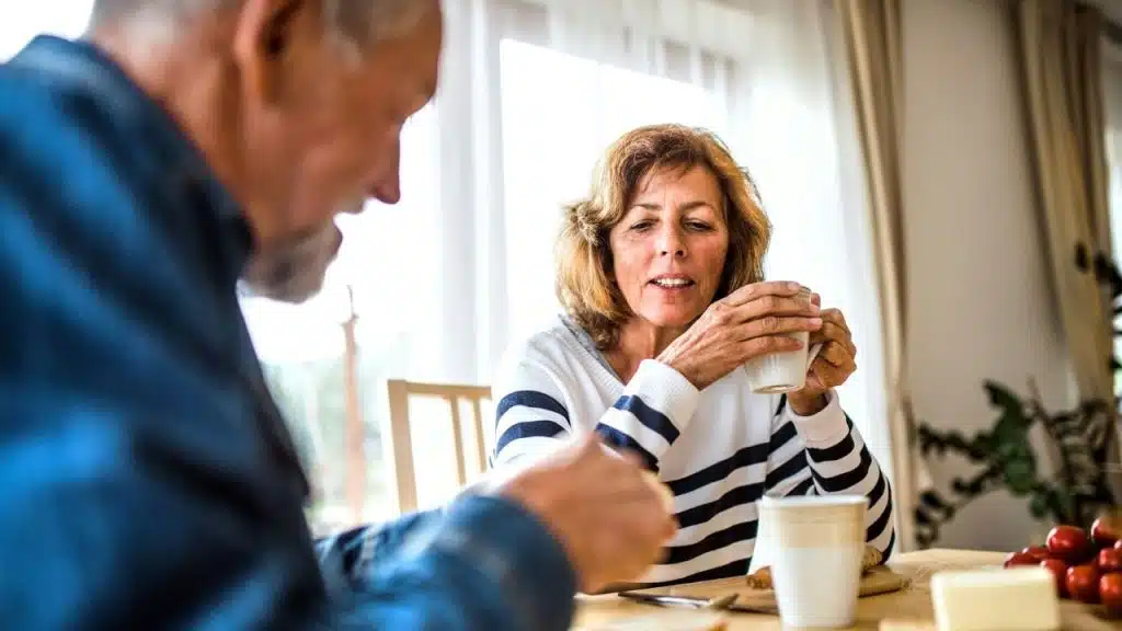 A mature woman holds a coffee mug while talking to her husband at a table.