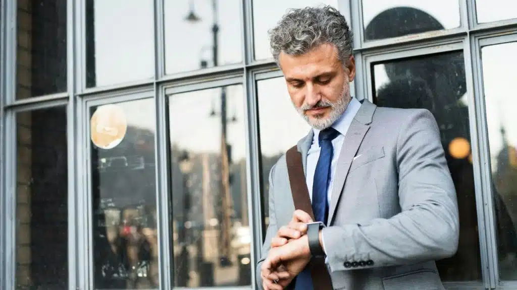 A serious, gray-haired businessman in a light suit checks his wristwatch outside a building.