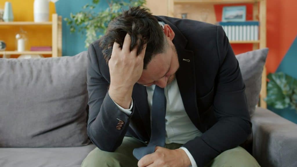 A distressed man in a suit and tie sits on a couch with his head in his hand.