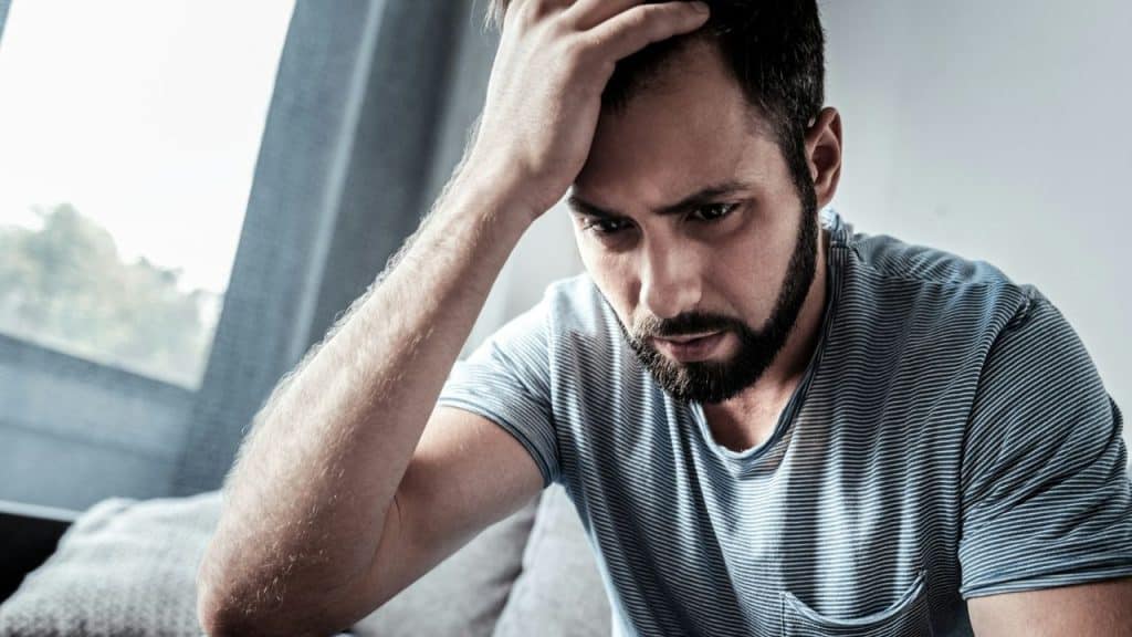 A bearded man in a striped t-shirt sits looking distressed with his hand on his head.