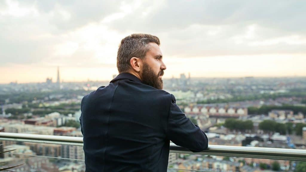 A bearded man in a dark suit stands on a high balcony, looking out over a city skyline.