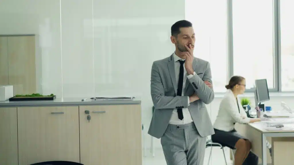 A businessman in a gray suit stands thoughtfully in an office while a colleague works in the background.