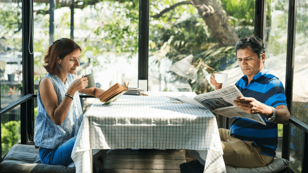 A man reads a newspaper and a woman reads a book while enjoying coffee outdoors.