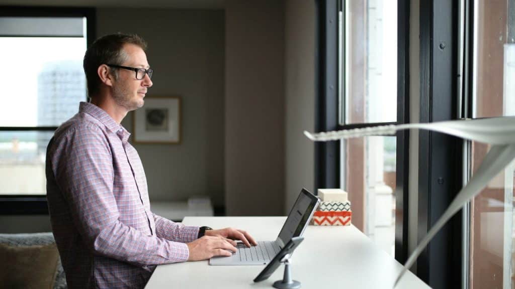 A bespectacled man in a plaid shirt stands at a desk, typing on a laptop and looking out the window.