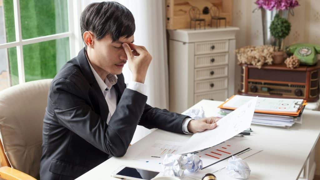 A man in a black suit sits at a desk, rubbing his eyes while reviewing papers and surrounded by crumpled paper.