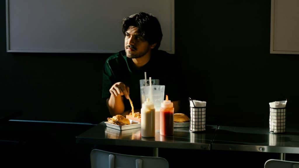A solitary man sits in the dark, eating french fries and a burger at a diner table.