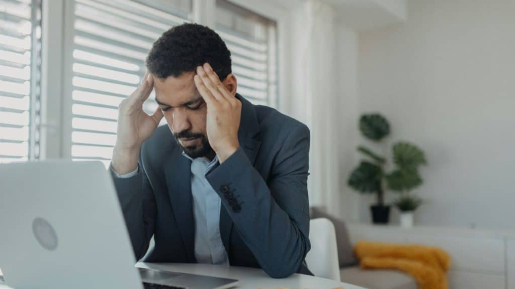 A frustrated businessman with a beard is seated at a laptop, holding his temples.