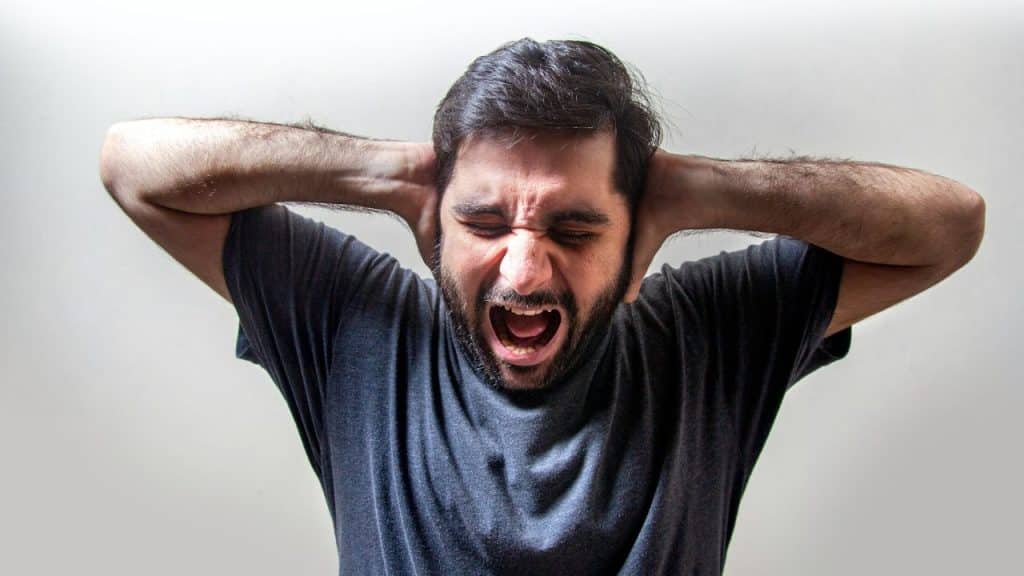 A frustrated, bearded man yells loudly, covering his ears with both hands.