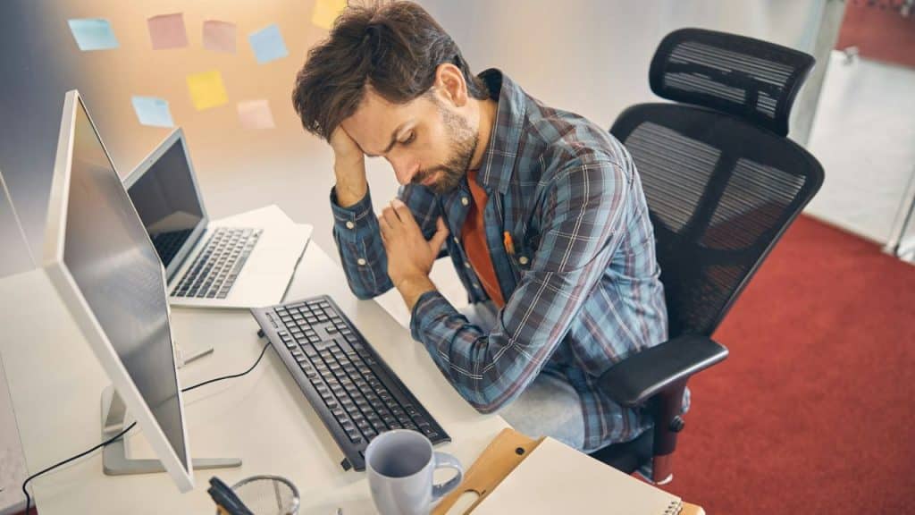 A weary man with a beard sits slumped at his office desk, resting his head on his hand.