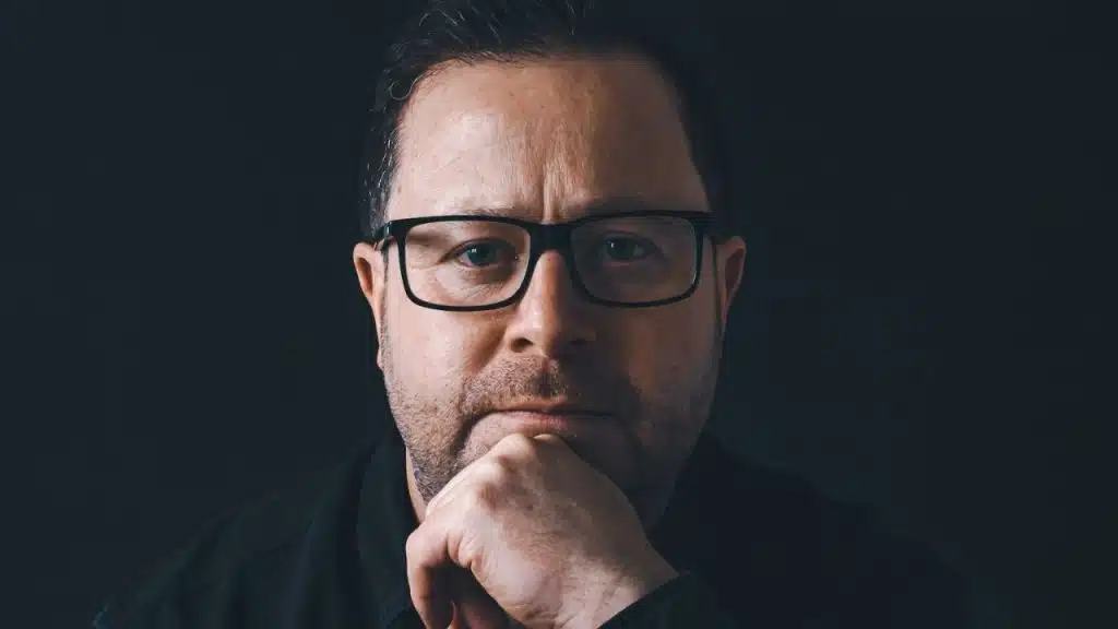 Man in black-rimmed glasses and a dark shirt, resting his chin on his hand.