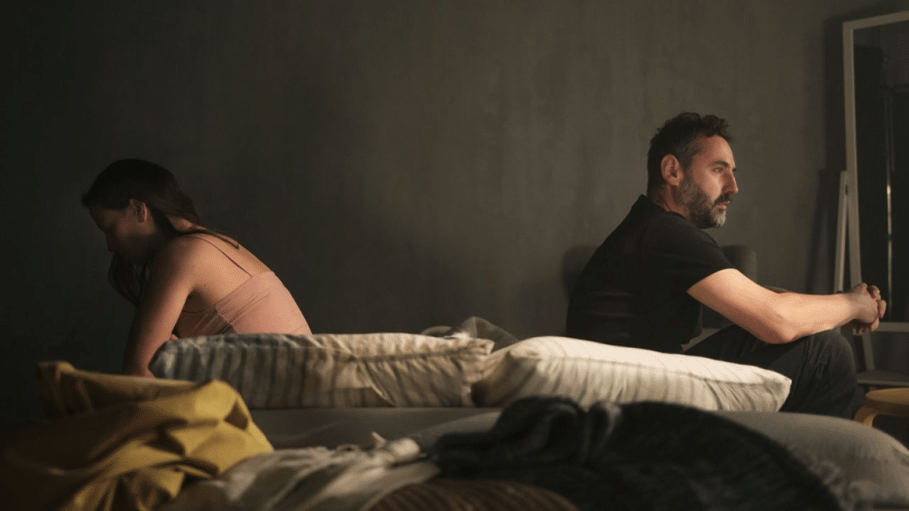 A couple sits on a bed, facing away from each other in a dimly lit, somber room.