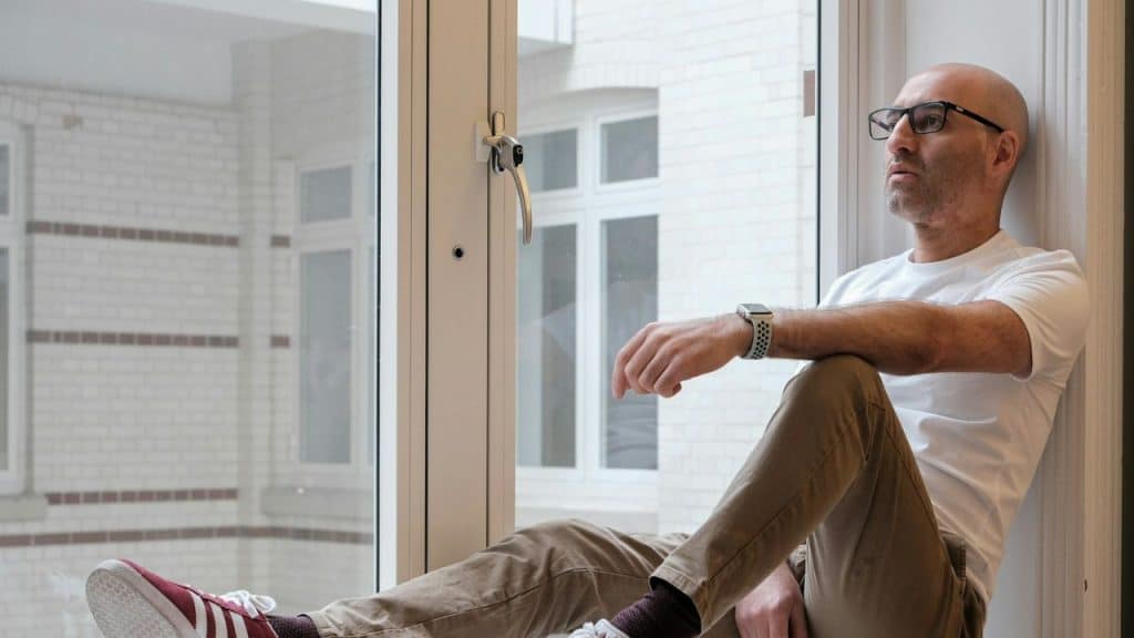 Bald man with glasses sits on a window ledge, looking out with a serious expression.