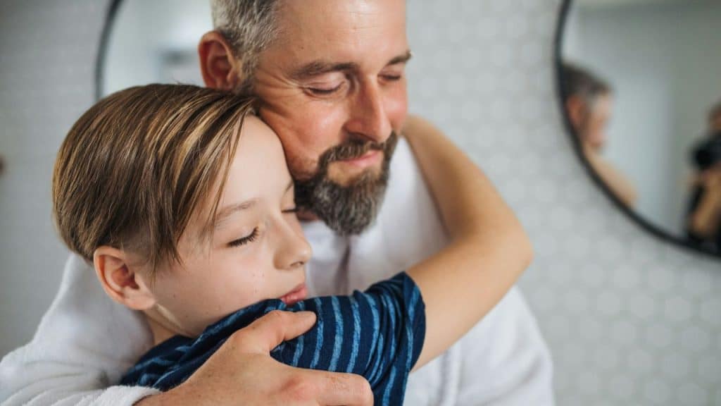 Man with a beard hugging a young boy, both smiling with their eyes closed.