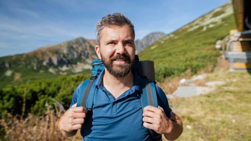 Smiling man with a beard and backpack wearing a blue shirt hiking in the mountains.