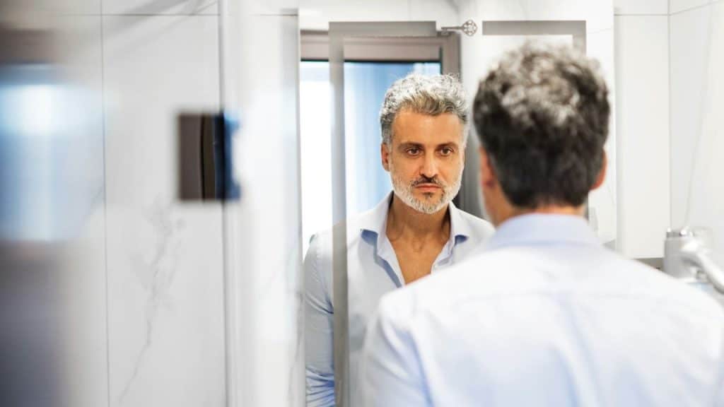 Man with gray hair and a beard, in a blue shirt, looking intently at his reflection.