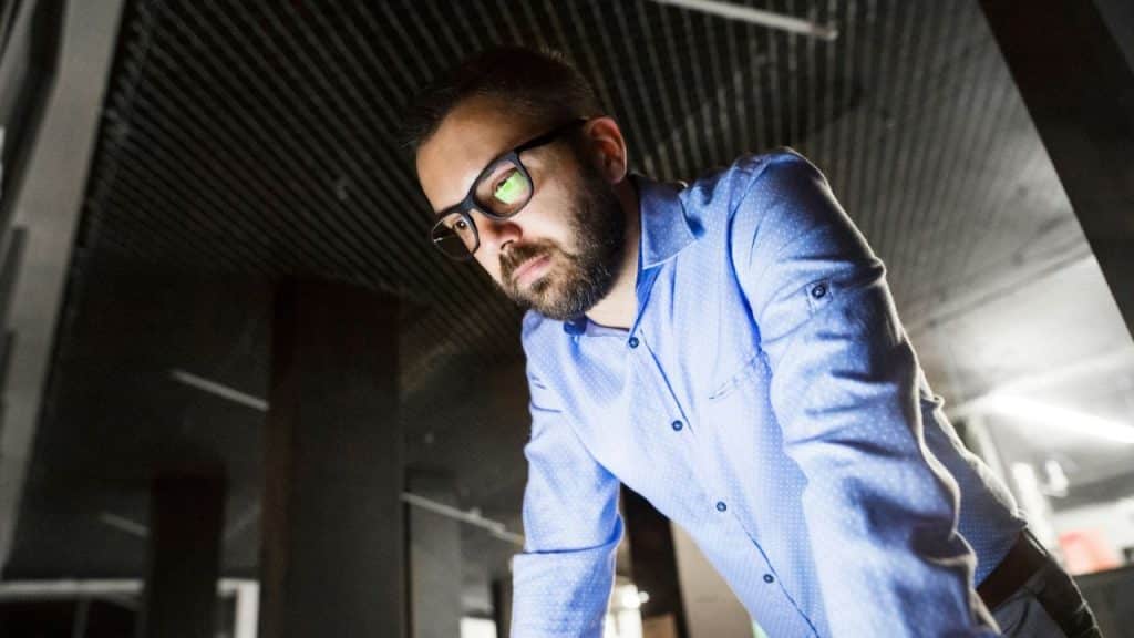 Man with a beard and glasses in a blue shirt looking down in a dark room.