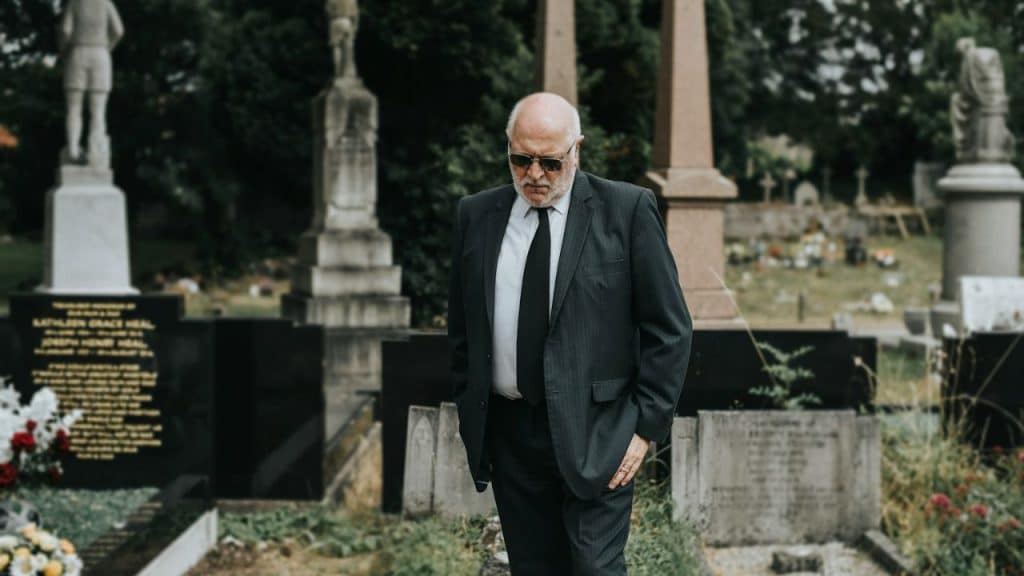 Older man in a dark suit and sunglasses walking thoughtfully through a cemetery.