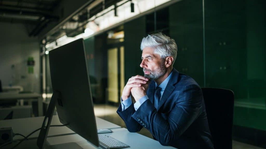 Man in a blue suit with gray hair and a beard, looking at a computer monitor at night.