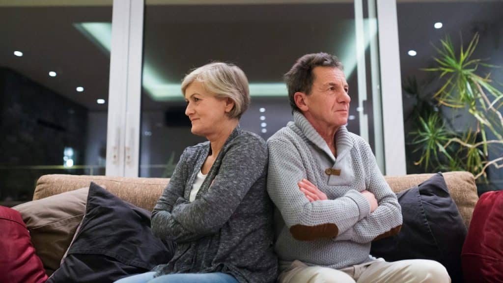 Middle-aged couple sitting on a couch back-to-back, arms crossed, looking away from each other.