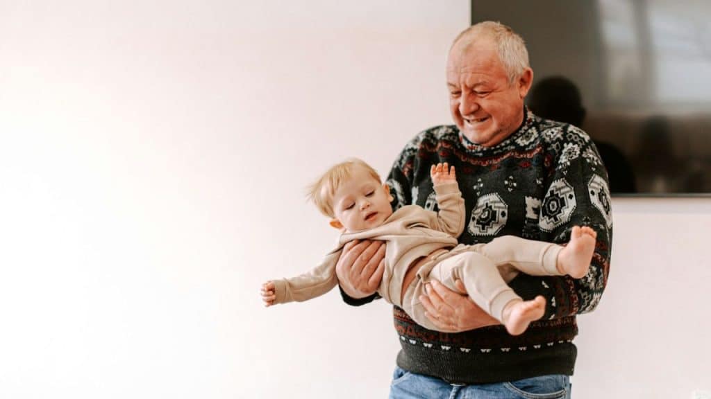Smiling older man holding and playfully swinging a toddler in a brightly lit room.