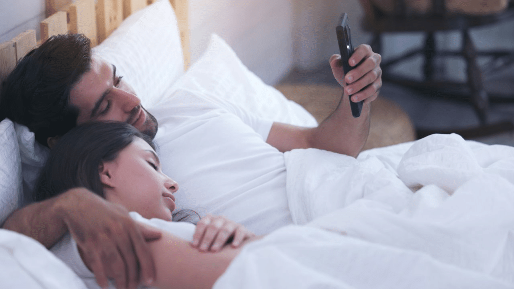 A smiling man holds and looks at a phone while cuddling a sleeping woman in bed.